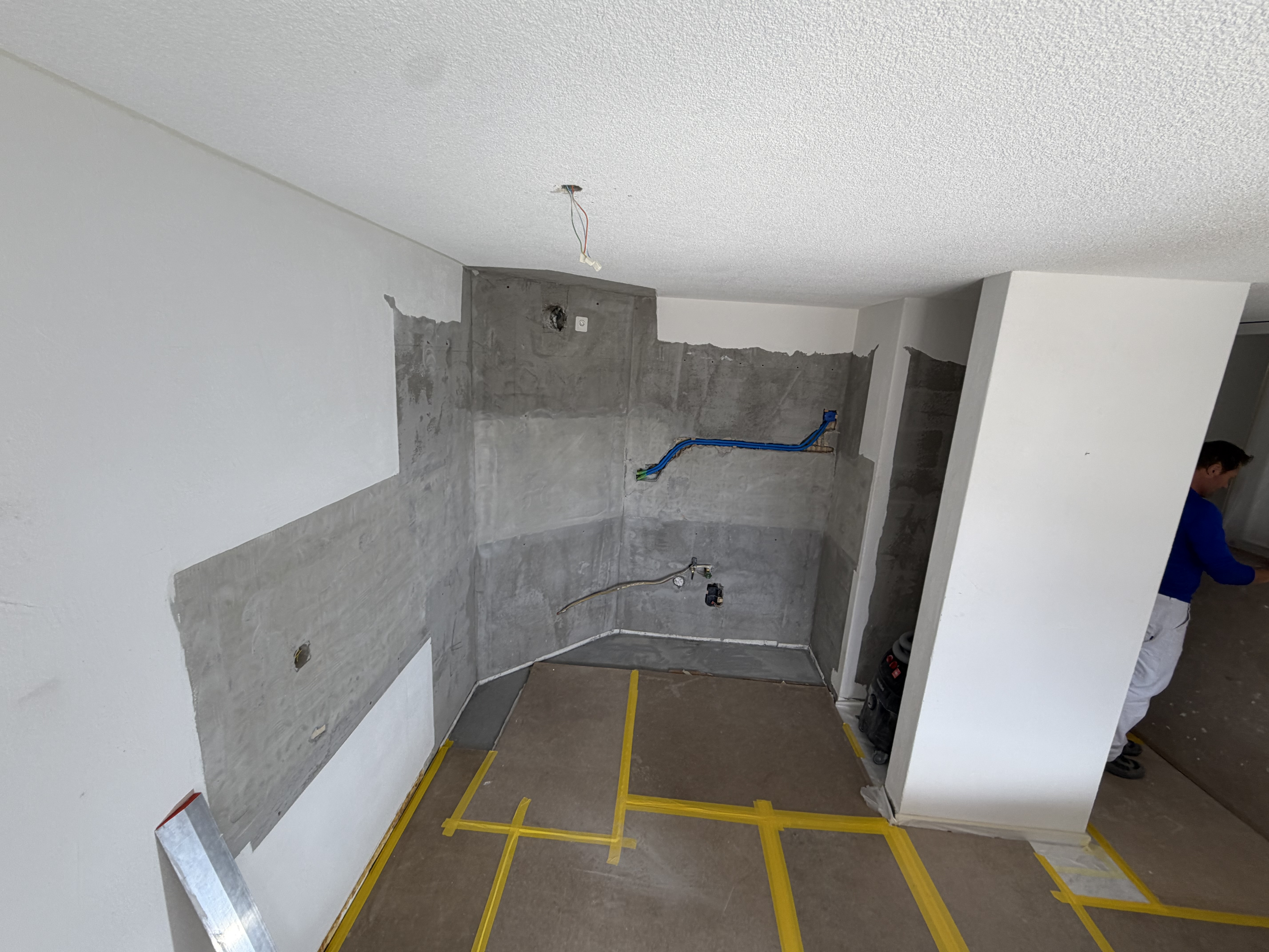 Renovationsbaustelle: freigelegte Betonwand mit Elektroinstallationen, sauber vorbereiteter Boden mit Klebeband, Handwerker im Hintergrund